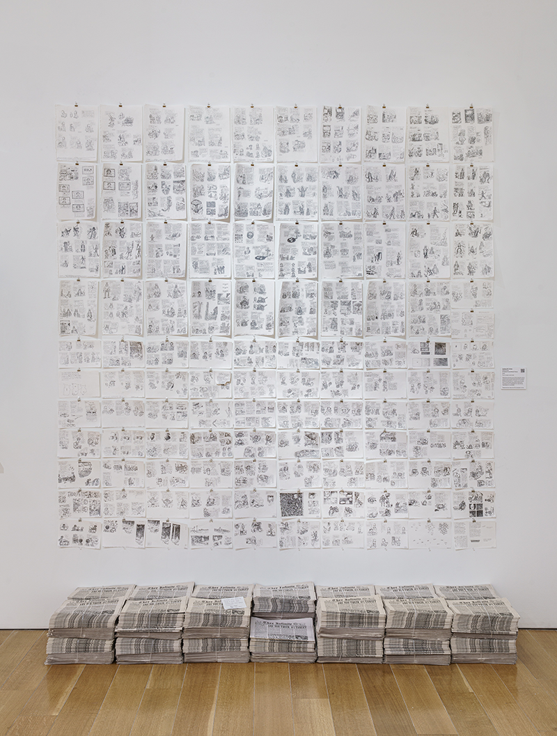 110 pencil drawings on paper in a grid on a wall with several stacks of newspaper comics below them